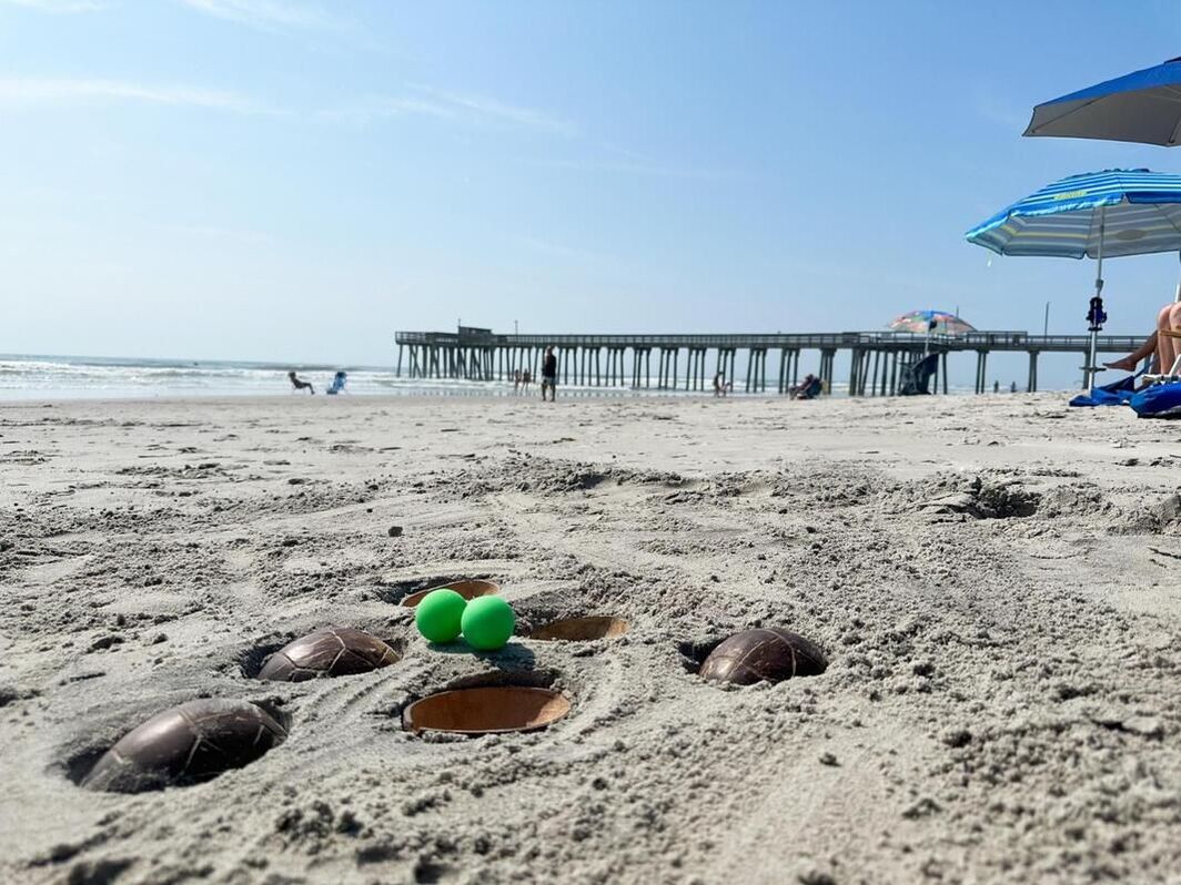 image of a beach with holes dug in the sand; 6 coconut shells are buried with 2 green balls sitting on the edge of a hole